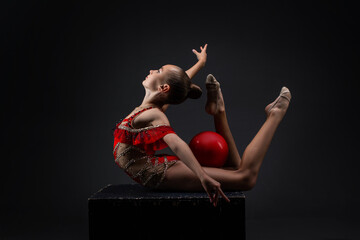 Young rhythmic gymnast girl posing in red leotard with red ball in dark studio