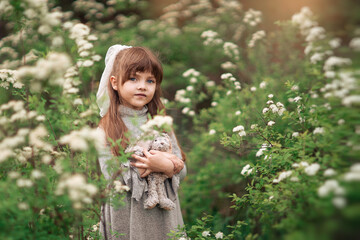 A cute little girl with brown hair holds favorite fluffy toy bunny among white flowers in the park. Background summer green flowering on the lawn. Child playing on the spring nature outdoors. Soft