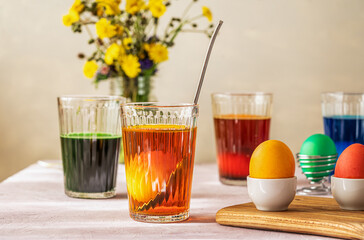 Process of painting Easter eggs. Four glasses with different colours, eggs inside and already painted, wild flowers