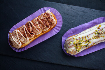 Two delicious eclair cakes - tiramisu and pistachio flavoured covered with cream, icing and chopped nuts, on a black background.