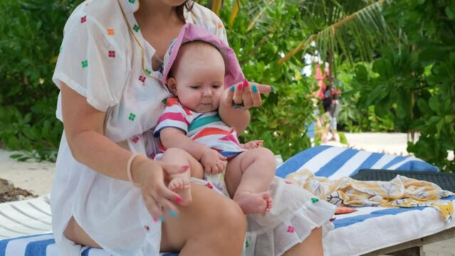 Young mother applying sunscreen protection cream on her baby while sitting on the beach chair on tropical island
