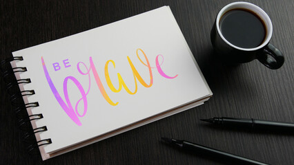  BE BRAVE colorful lettering in notebook with cup of espresso and pen on black wooden desk