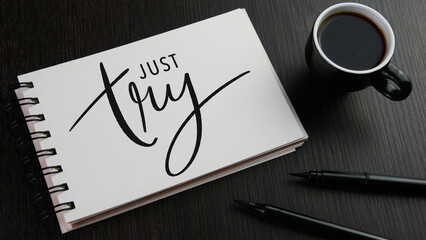 JUST TRY black hand lettering in notebook with cup of espresso and pens on black wooden desk