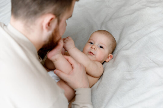 A young father with a beard is kissing the foot of his newborn daughter, who lying on the bed.