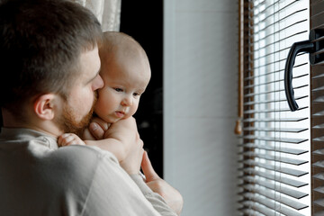 A young father with a beard is holding a newborn daughter in his arms near a window with wooden blinds.