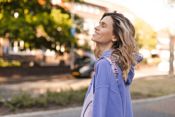 Attractive curly blonde woman walk on the city park street. Girl wear purple hoodie, pink bag and look happy and smiles. Woman go on the street, look from back, she turn.