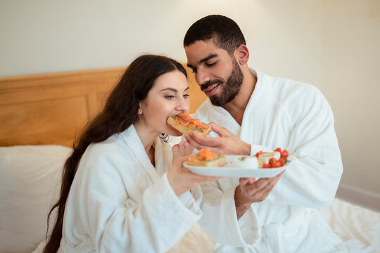 Husband Feeding Wife Giving Her Sandwich Having Breakfast In Bedroom - Powered by Adobe