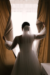 the bride in a luxurious dress stands near the window and opens the curtains