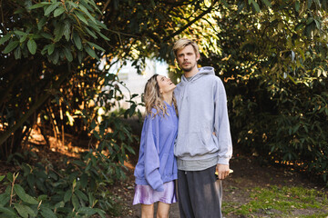 Fototapeta premium Couple walk outdoors in the park. Woman warped man and look at him laughing. Girl wear purple hoody and skirt, man wear grey hoody and pants. Fashion outfit of couple, love relationship.