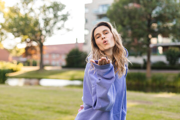 Attractive curly blonde woman walk on the city park street. Girl wear purple hoodie, pink bag and look happy and smiles. Woman send blow kiss while go on the street, look at camera.