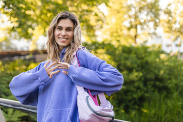 Attractive curly blonde woman walk on the city park street. Girl wear purple hoodie, pink bag and look happy and smiles. Woman walk on the street, close up portrait of woman show her smile.