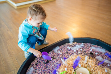 Little girl playing with sensory colorful rice. Sensory development and experiences, themed activities with children, fine motor skills development - obrazy, fototapety, plakaty