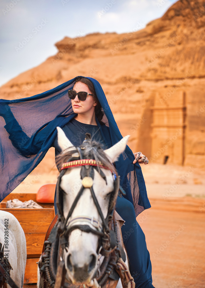 Wall mural Young woman with dark long dress, head scarf and sunglasses, sitting on a horse, blurred Hegra or Mada’in Salih ruins in background - female tourist travel influencer like photo from Saudi Arabia