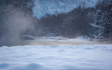 Cold winter river, steam visible above water, dark trees on both sides, focus to fresh snow in foreground