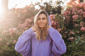 Smiling curly blonde hair girl puts on a hood with purple hoodie, walking on the street with...