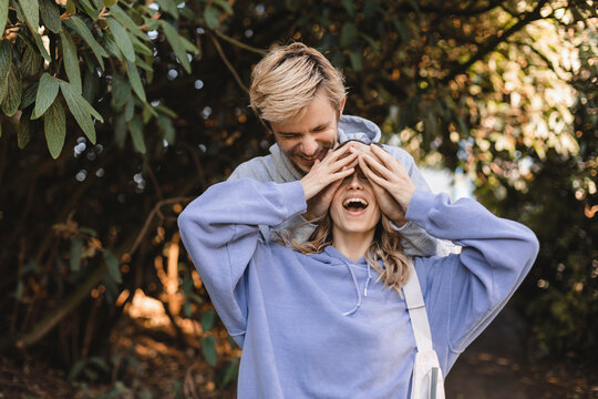 Photo of funny couple in love walking guy palms cover lady eyes wear casual clothes garden park outdoors. Charming couple hiding eyes outside in park. Man and woman wear hoody.