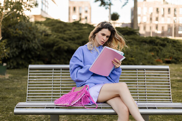 Blonde curly woman sitting on bench and look at her notebook, read it, girl wear purple hoody, skirt and pink tote bag. Woman study in the park or read her diary, notes, dreaming and think about life.