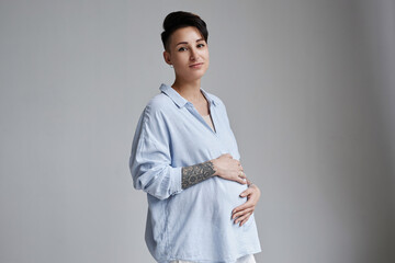 Minimal portrait of pregnant young woman with tattoos posing against grey background and looking at camera, copy space