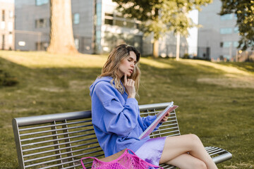 Blonde curly woman sitting on bench and look at side, thinking, hold finger near chin, search decision or problem solution. Woman study in the park or read her diary, notes.