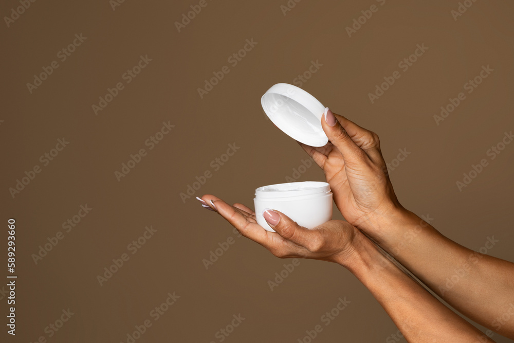 Wall mural African american woman holding jar with facial cream and opening it, posing over brown background, free space, closeup