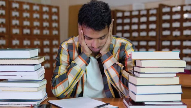 Tired indian student, in casual clothes, sits at a desk in a library between books, holds his hands on his head, looks tired of reading