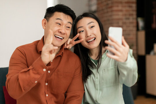 Excited asian couple taking selfie, gesturing peace signs, taking pictures for happy memory, social media or blog post - Powered by Adobe