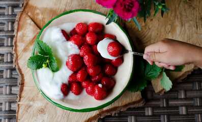 Spoon holding in child hand and red ripe fruits with sweet cream in the bowl. Ecological natural dessert as a healthy food. Top view. Summer season for fruits.