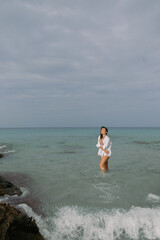 a young girl with wet hair in a wet white shirt is standing in the water on the seashore by the rocks