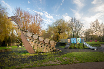 Wooden boat climbing frame structure in outdoor natural public playground with slide in background.