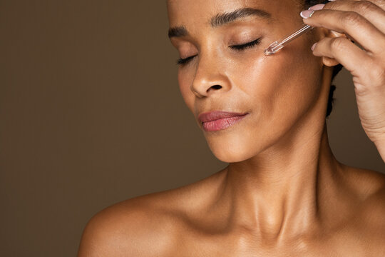 Closeup shot of black middle aged woman applying facial serum moisturizing skin, brown background, free space