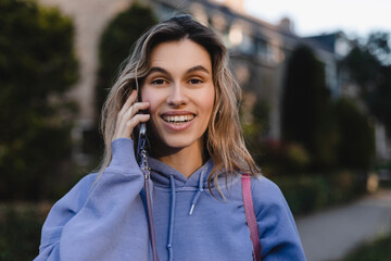 Happy blonde curly woman talking phone and look happy, smiling. Portrait of gorgeous smiling female using mobile phone. Girl wear purple hoody and pink bag.