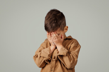Sad caucasian 6 years old little kid covering face with hands, crying isolated on gray studio...
