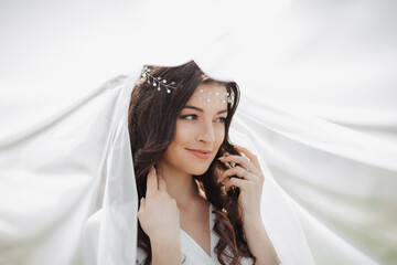 A beautiful bride with long curly hair and a delicate tiara on her head, wrapped in a white veil,...