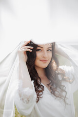 A beautiful bride with long curly hair and a delicate tiara on her head, wrapped in a white veil, gently holds her hair. Portrait of a girl looking into the lens. Natural makeup