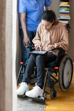 Woman with disability using tablet PC while sitting on wheelchair by caregiver at home
