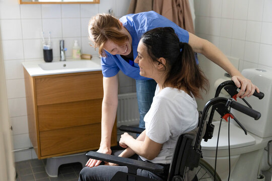 Happy female caregiver talking to woman with paraplegia in bathroom at home