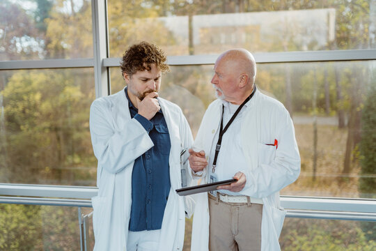 Senior male doctor holding digital tablet discussing with colleague while standing against window at hospital