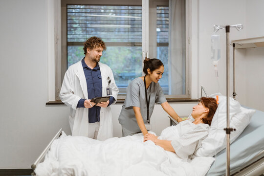 Smiling female nurse talking to senior patient lying on bed by doctor in hospital