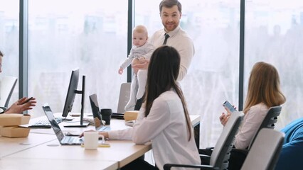 Man is standing, talking to empoyees and holding little baby boy in hands. Group of people working in the modern office together.