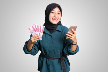 Portrait of excited Asian hijab woman in casual outfit showing one hundred thousand rupiah while...
