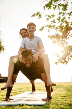 Father carrying children on back while practicing push-ups in park during sunset