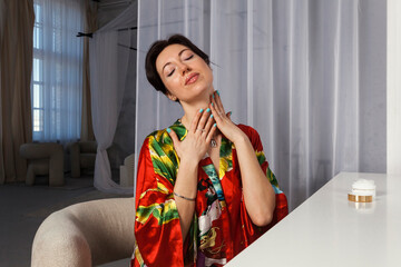 A happy woman in a sensual bathrobe using and applying a cream on a face and body after bath.