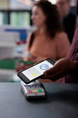 Young adult putting smartphone on pos terminal using contactless to pay for health care treatment in pharmacy, shopping for pain pills. Clients waiting in line at drugstore counter. Close up