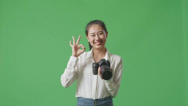 Asian Photographer Looking At The Pictures In The Camera And Showing Okay Gesture While Standing On Green Screen Background In The Studio
