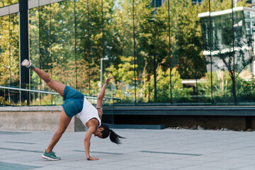 mujer morena con ropa urbana practicando capoeira en la ciudad y haciendo flexiones. - obrazy, fototapety, plakaty