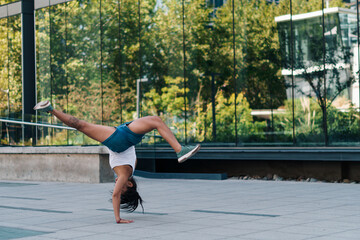 mujer morena con ropa urbana practicando capoeira en la ciudad y haciendo flexiones. - obrazy, fototapety, plakaty