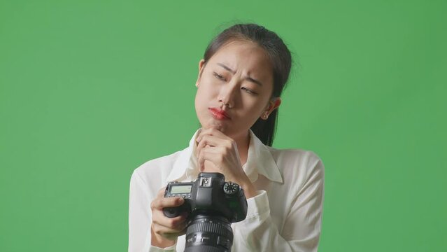 Close Up Of Asian Photographer Thinking About The Pictures, Footage And Raising Her Index Finger While Looking At The Pictures In The Camera On Green Screen Background In The Studio
