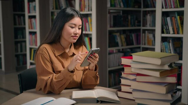 Smiling asian woman student in university library check timetable using mobile application on phone chinese girl chatting in social media on cellphone search information online take break in learning