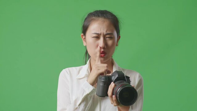 Close Up Of Asian Photographer Using A Camera Taking Pictures And Making Shh Gesture While Standing On Green Screen Background In The Studio
