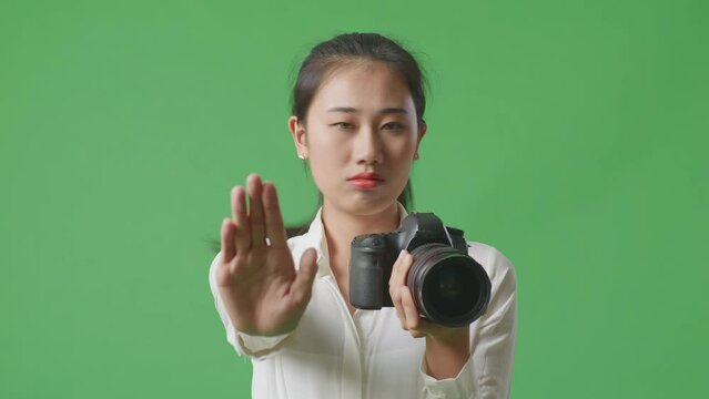 Close Up Of Asian Photographer Using A Camera Taking Pictures And Making Stop Hand Sign While Standing On Green Screen Background In The Studio

